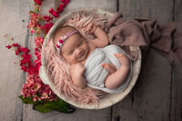 Newborn baby girl wrapped in white with a pink floral headband, sleeping in a rustic bowl on pink fur at Kara Reese Photography in Waukesha, WI.
