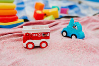 A small red toy fire truck and a blue toy vehicle are placed on a patterned red blanket, with colorful blocks and a stacking toy in the blurred background.