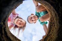 Two children leaning over and smiling as they look down through a circular coquina rock hole at Palm Coast Beach during a beach child portrait session in Palm Coast. (Photographed by Nature's Child Photography)