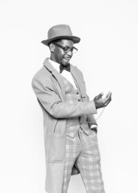 Luxury fine art portrait from Luxe Portraiture LV featuring a man in a plaid three‑piece suit, bow tie, overcoat, and fedora, holding a pocket watch in a classic black‑and‑white vintage style photograph that evokes early 20th‑century fashion and refined elegance.