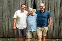 Three adult men standing together in front of a weathered wooden barn during an extended family session