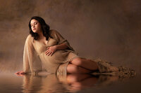 Pregnant woman in a soft, draped white gown cradling her belly while seated on a floor in a bright Waukesha studio, showcasing wardrobe options provided by the photographer.