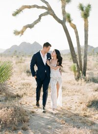 Bride Sophie with long, sleek hair, a Kohnur romantic wedding hairstyle, and radiant soft glam bridal beauty makeup, walking with groom Joshua in the sunny desert near Joshua Trees.