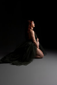 Expecting mother with long red hair wearing a dramatic dark green maternity gown while kneeling and gently holding her baby bump during an artistic studio maternity portrait on a dark background photographed by Inland Empire maternity photographer Kaitlyn Dawn Photography.