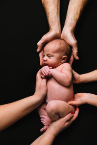 A naked baby lies on a black backdrop with her parents' and sibling's hands laying against her.