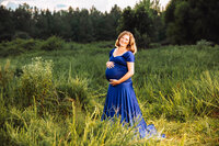 A redheaded pregnant woman in a royal blue dress stands in a large overgrown field on a cloudy day. The landscape is vast.
