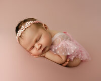 Sleeping newborn baby girl wearing a delicate pearl headband and pink lace outfit, posed on a soft blush backdrop during a studio newborn photography session by Kaitlyn Dawn Photography.