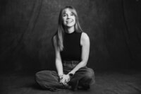 A black and white picture of a blonde woman with straight hair sitting cross-legged on a dark gray backdrop. She wears jeans and a black tank top, smiling brightly.