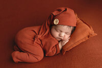 Newborn boy wearing a burnt orange onesie with matching sleepy cap during his newborn photography session at Jennifer Brandes Photography.