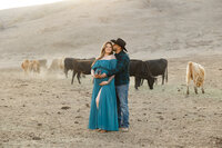 Expecting couple standing together in a rustic pasture with cattle during an outdoor maternity photography session while the father kisses the mother’s head photographed by Kaitlyn Dawn Photography.