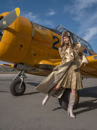 Model in layered gold and beige fashion standing beside a yellow vintage training aircraft.