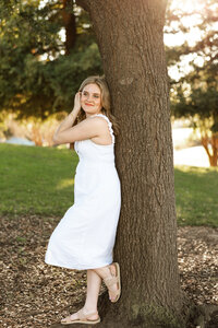 High school senior girl standing beside a tree in a sunlit park wearing a white dress, outdoor senior portrait by Kaitlyn Dawn Photography.