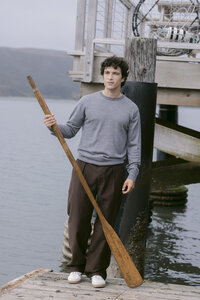 Model in a grey merino crewneck and brown wide-leg trousers holding an oar on the dock