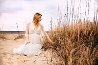 A pregnant woman caresses the beach grass. She sits on golden sand with a cloudy sky above her.