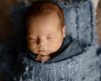 Close-up portrait of newborn baby boy posed in a wicker basket with a soft blue wrap on a rustic wooden backdrop during a studio newborn photography session by Kaitlyn Dawn Photography in Yucaipa.