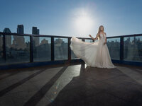 A woman in a flowing white gown stands on a rooftop terrace at sunset, city buildings behind her, sunlight streaming and casting long shadows—captured by a creative portrait photography Washington DC specialist.