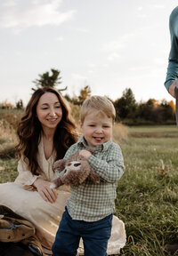 A heartwarming family photo capturing a baby boy running towards the camera with a big smile while mom crouches in the background in a cream dress—joyful, candid lifestyle photography.