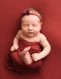 newborn baby girl with headband on burgundy fabric swaddled