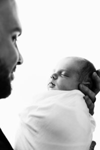 A black and white backlit picture of a newborn baby in her father's hands. The dad is slightly out of focus off to the side smiling down at her.