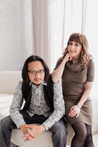 A man with long hair and glasses sits on a sofa, hands clasped, wearing a patterned shirt and vest. A woman in a sweater dress smiles beside him, her hand resting on his shoulder—an editorial portrait photography Washington DC metro style.