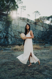 A woman in white linen walks barefoot at at rock quarry at sunset holding a ceramic vessel