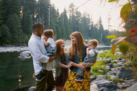 colorful family portrait of a mom in a yellow dress with red hair, her two boys in overalls and her daughter with red hair and dad in button down shirt all looking at each other in front of a river