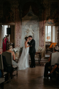 Bride and groom share their first kiss during intimate Italy destination wedding ceremony in ornate venue.