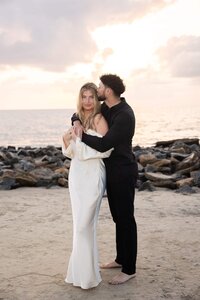 A couple embraces on the beach at sunrise during their engagement session. She wears an off-the-shoulder ivory gown, and he’s in an all-black outfit. The soft morning light and rocky shoreline create a serene, intimate backdrop. Captured in a natural, documentary style by Phavy Photography—a St. Augustine engagement photographer known for luxury beach sessions that feel effortless and timeless.3