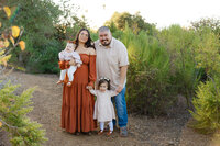 Mother and father standing together outdoors holding their two young children surrounded by greenery, natural light family portrait by Kaitlyn Dawn Photography.