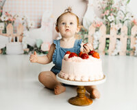 Baby smiling and playing with their strawberry cake during a fun studio cake smash session. Photographed by Yucaipa baby photographer Kaitlyn Dawn Photography.