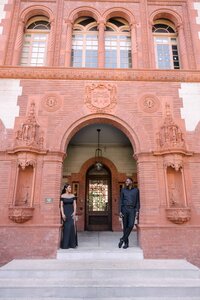 A fashionable couple poses in front of the grand, historic architecture of Flagler College in St. Augustine, Florida. She wears an elegant off-the-shoulder black gown, and he’s dressed in a classic all-black ensemble. Captured by Phavy Photography, a St. Augustine engagement photographer known for crafting luxury, editorial-style engagement sessions in iconic locations.
