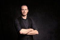 A dark haired man with light blue eyes holds a serious expression with his arms crossed in front of him. He wears a gray shirt with a black vest and stands in front of a gray backdrop. The lighting is dramatic.