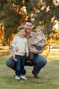 Father kneeling in a grassy park holding his two young sons during golden hour, warm natural light family portrait captured by Kaitlyn Dawn Photography, Yucaipa professional outdoor family photographer.