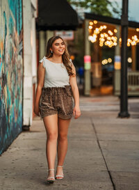 Senior portrait in downtown Mobile, Alabama — walking on a sidewalk near a grafitti wall with lights glimmering in the background.