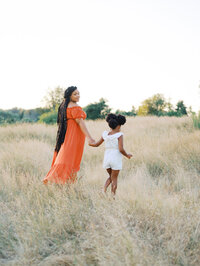 Mom and daughter holding hands and walking in a grassy field in Discovery Park, mom looking back at camera