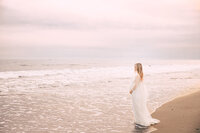 A blonde pregnant woman stares out into the ocean. She stands just in the water's edge with her side to the camera. The sky is blue with hints of pink.