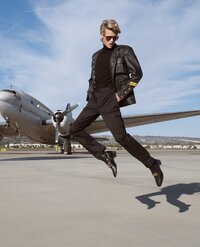 A person wearing a black leather jacket, black pants, and sunglasses is mid-jump in front of a vintage propeller airplane—captured in the bold style of a cinematic portrait photographer Washington DC, under blue skies with wispy clouds.