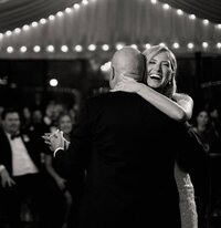 Bride embraces her father during a first dance at The Club at 12 Oaks, guests watching beneath draped café lights.