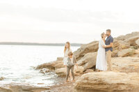 A lake family photo as mom and dad stand together on the rocks while the children are playing in the water.
