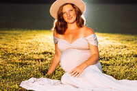 A pregnant woman in a cream fedora sits on short cut grass. It is dusk and she is backlit with small fireflies glistening around her.