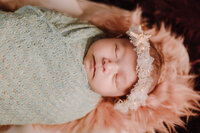 A newborn baby sleeps peacefully in a seafoam green wrap and beachy headband. She sleeps on a coral colored fur within a large shell.