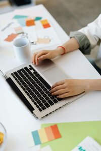 A person is typing on a laptop at a white desk, which is covered with colorful swatches, design papers, and a white coffee mug.