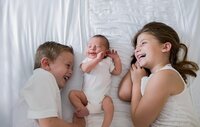 St. Johns newborn capturing a joyful sibling moment as a baby smiles between big brother and sister, their faces close and laughter shared in soft natural light. - A portrait experience by Nature's Child Photography.