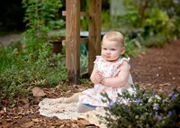 Baby girl in a floral dress sitting on a knit blanket among garden plants with a serious expression during an outdoor child in Mandarin. - A portrait experience by Nature's Child Photography.