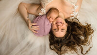 A mother lays on a tulle dress smiling up brightly while holding her wrapped newborn against her face.