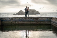 reflection of a couple embracing on a wall surrounded by water and the ocean at sunset