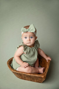 Baby girl in green romper posed in a wooden heart bowl.