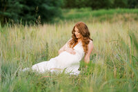 Mom with auburn hair and white dress lays in grass and looks at belly during maternity photo session