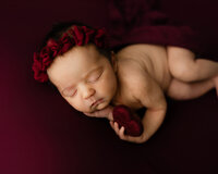 Sleeping newborn baby girl holding a small red heart prop while wearing a burgundy floral headband during a studio newborn photography session in Yucaipa by Kaitlyn Dawn Photography.