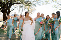 Bride walking and laughing with her bridesmaids in light blue dresses while holding colorful bouquets on the lawn at Burritt on the Mountain in Huntsville, Alabama, photographed by Perry Hancock.